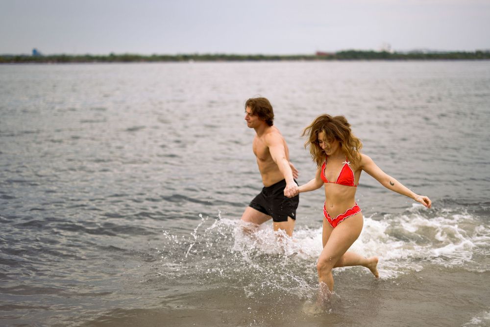Two people in swimwear splash and play in shallow water at a lake on an overcast day.