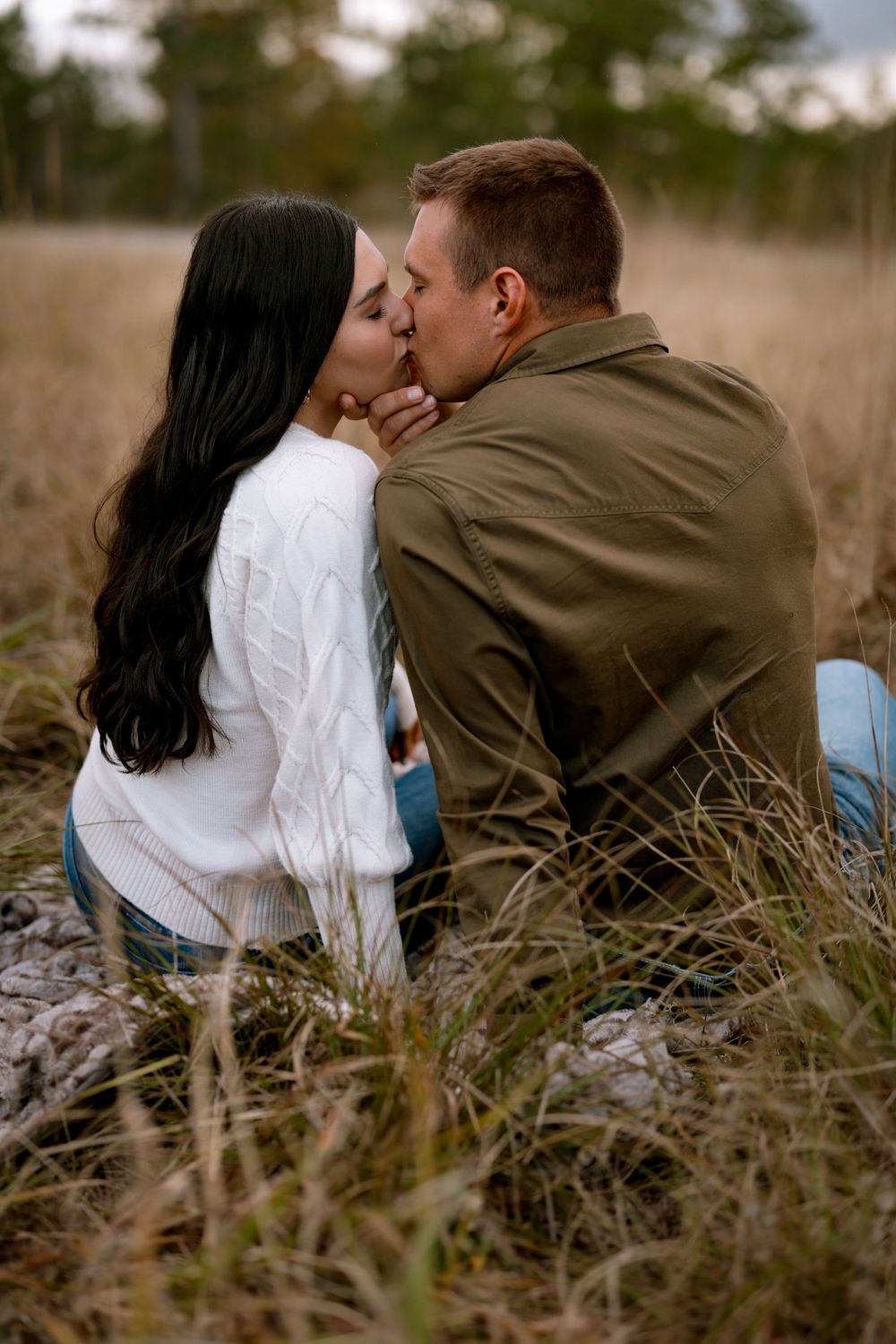 A romantic couple shares intimate moments together while sitting in tall grass during a photo session at golden hour.