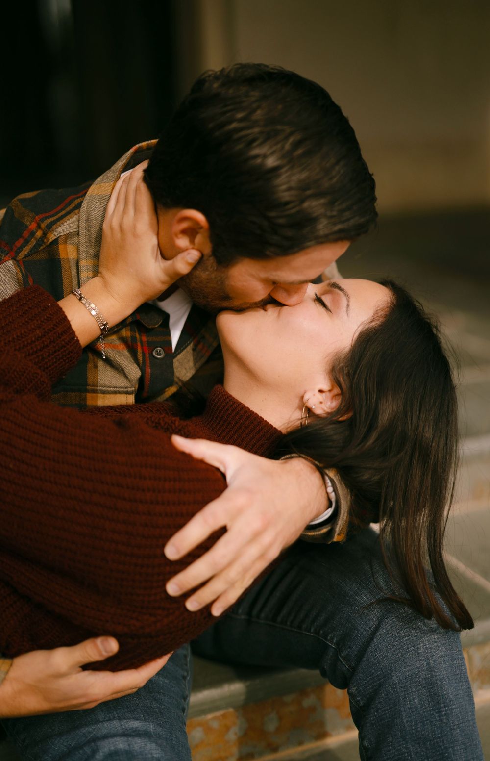 A romantic couple in warm sweaters share a tender kiss while embracing in moody lighting.