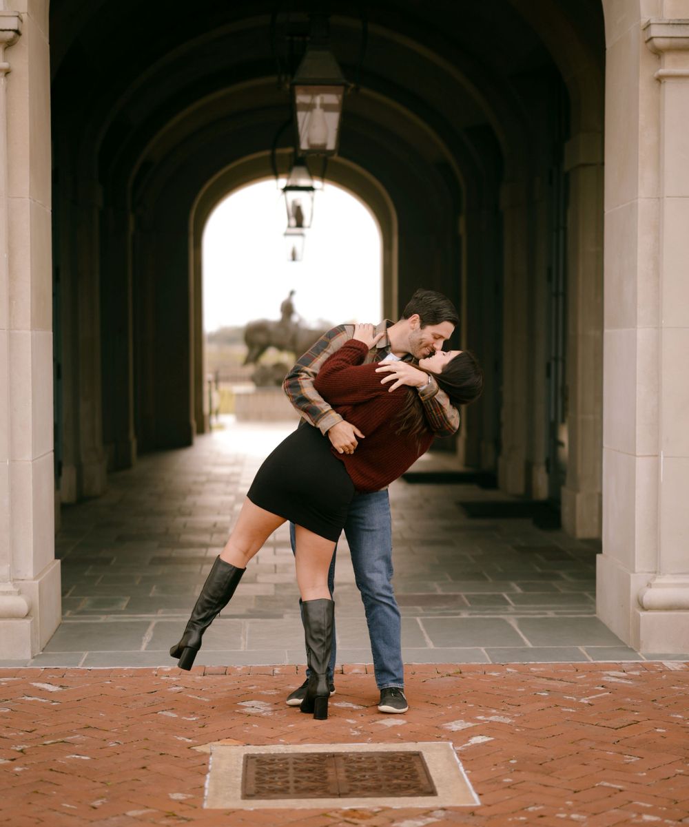 A romantic couple embraces in a dip pose under an arched passageway with stone columns and brick flooring.