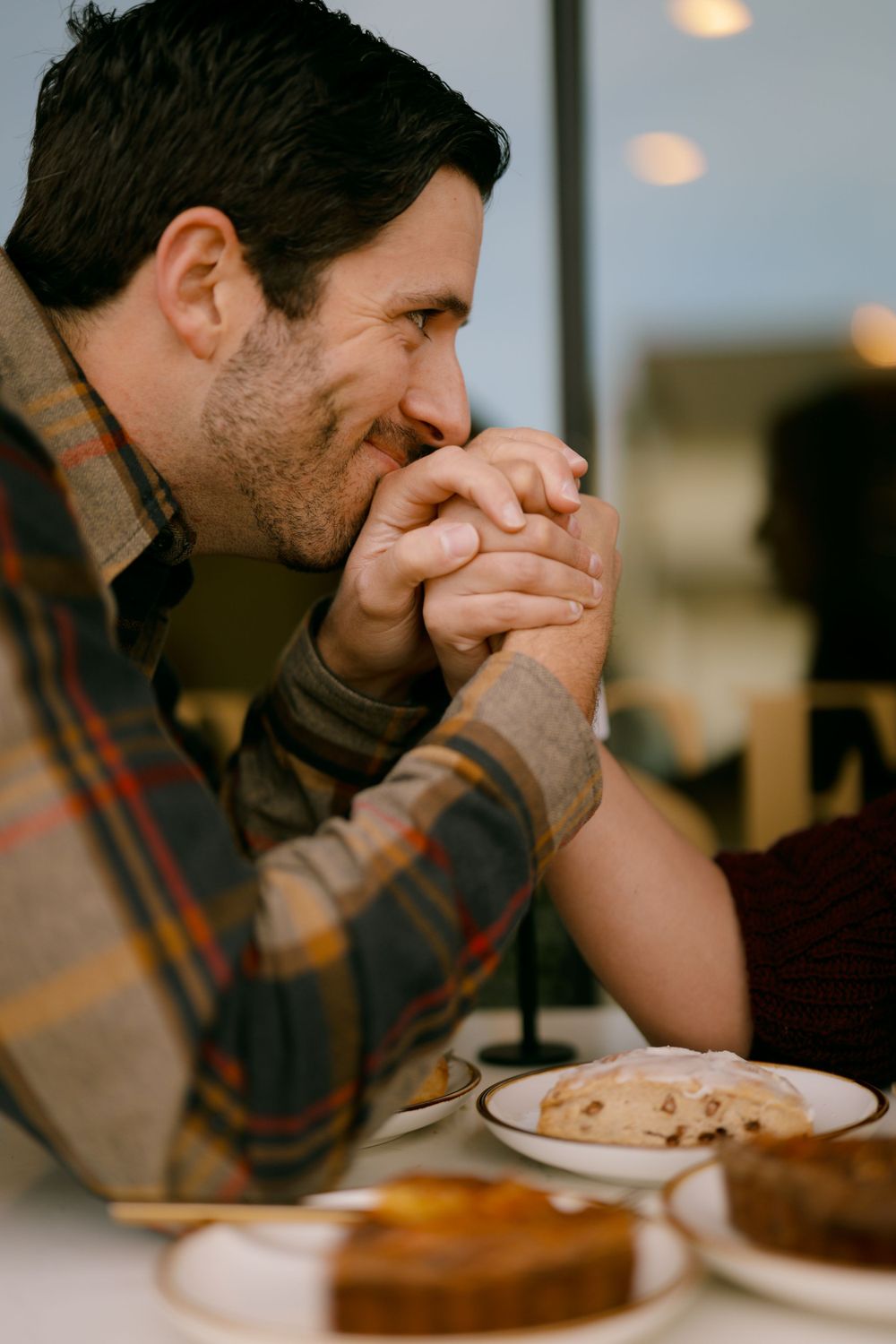 Person wearing plaid shirt sits at restaurant table with hands clasped near face in contemplative pose.