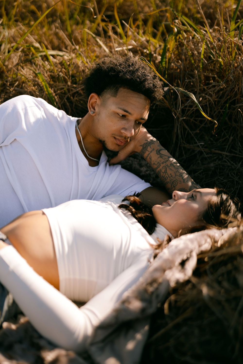 A tender maternity photoshoot outdoors with a couple lounging in tall grass at golden hour.