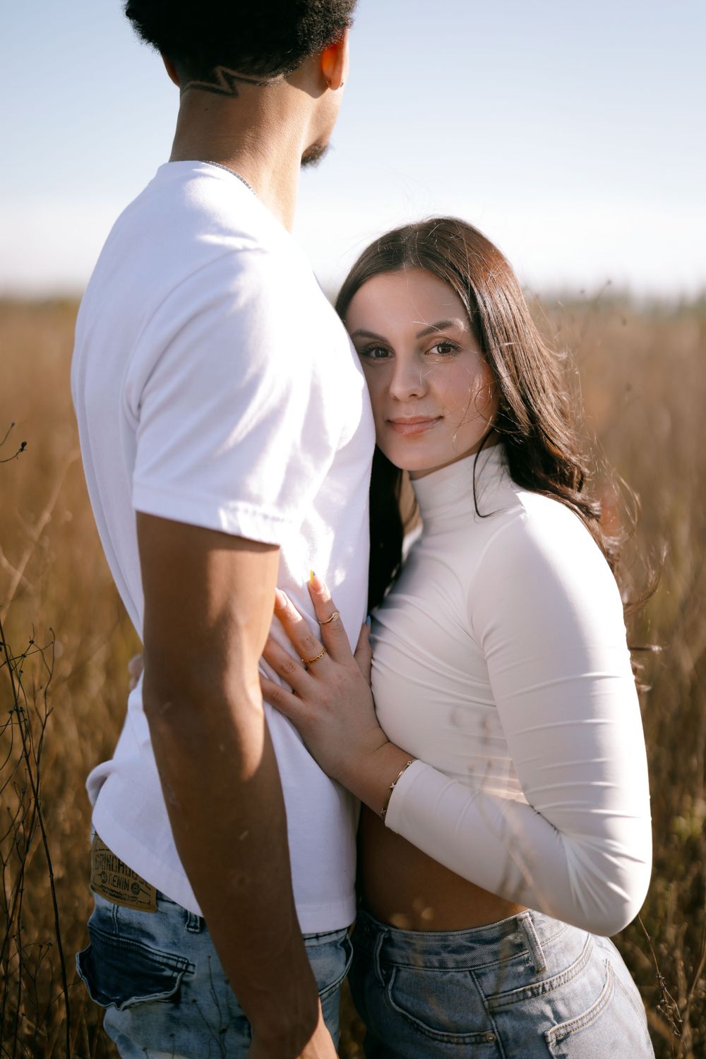 A couple in white tops and jeans embrace tenderly while standing in a sunlit field of tall grass.