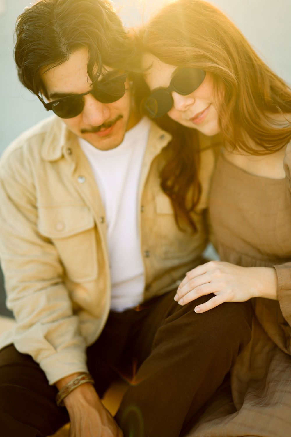Two people in sunglasses sit close together outside in warm sunlight with a dreamy vintage film aesthetic.