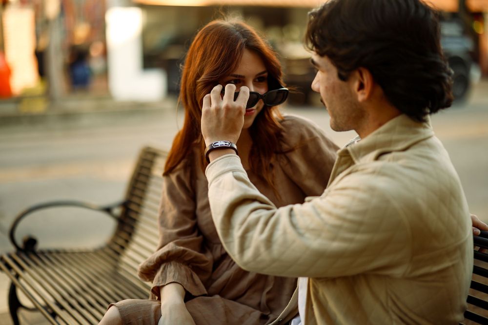 Two people share a tender moment on a bench as one takes a photo of the other with a vintage camera.
