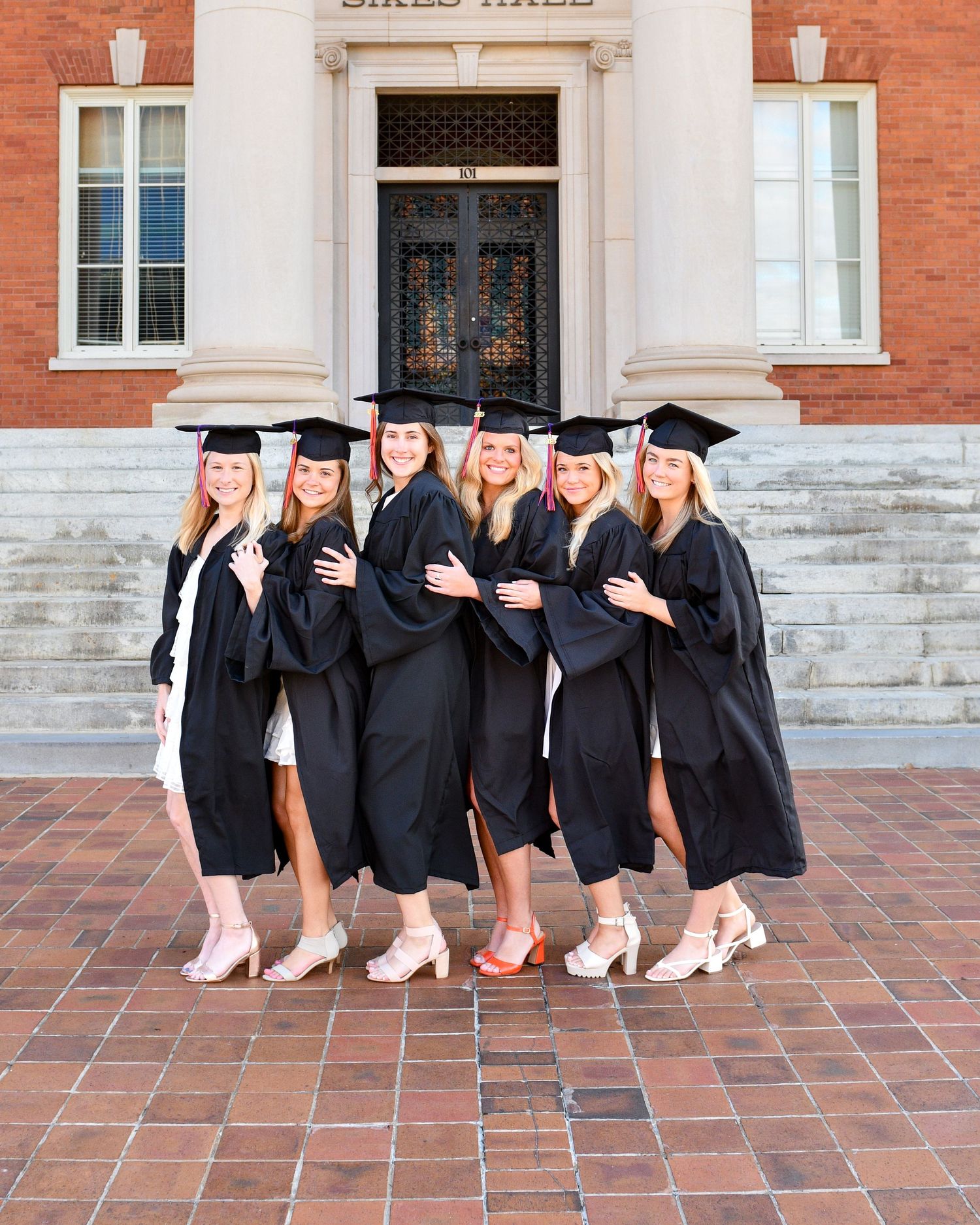 Group of graduates in black caps and gowns posing on steps of brick building.