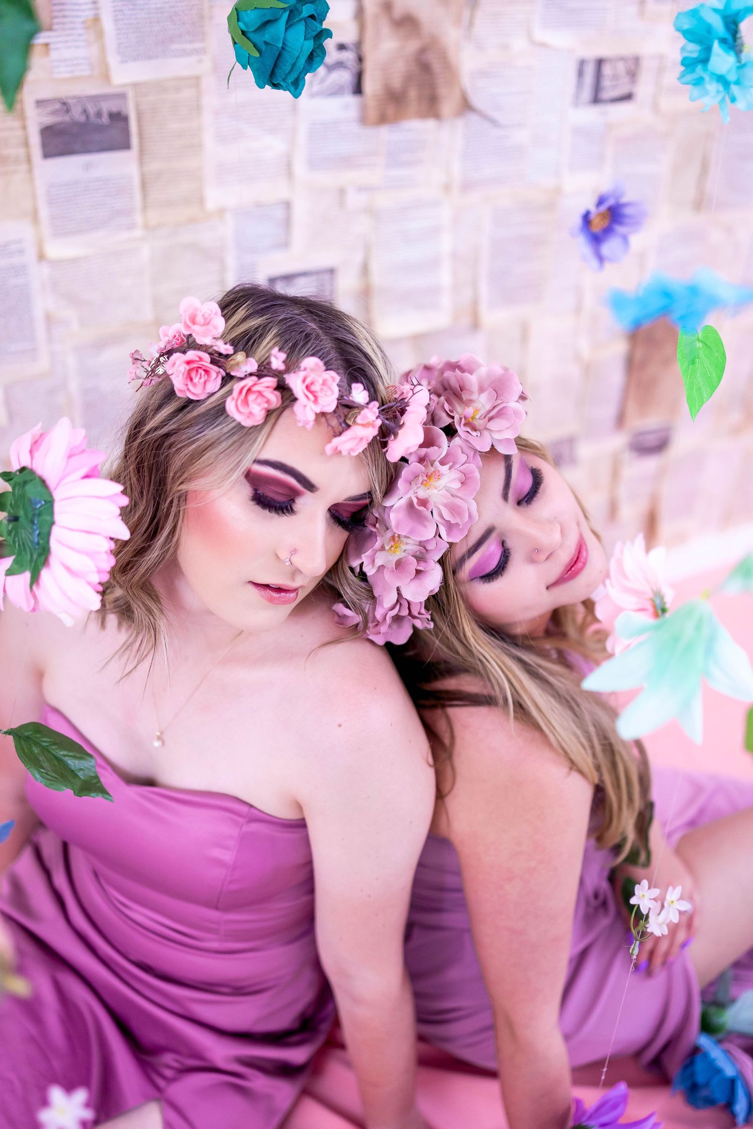 Two models wearing purple dresses and pink floral crowns pose artistically against a white backdrop with blue accents.