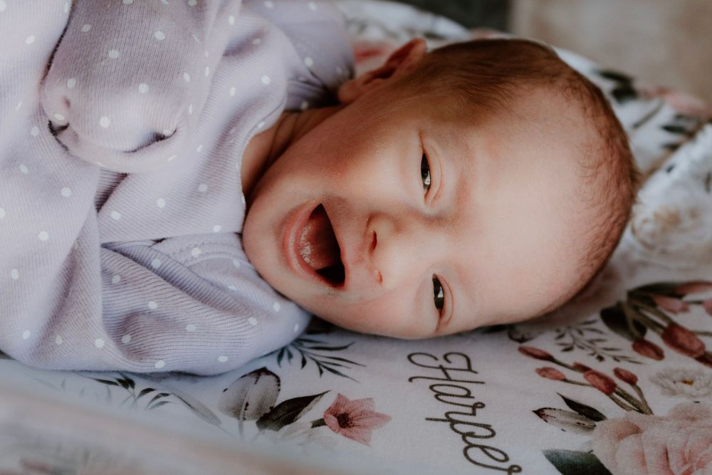 Smiling newborn baby in lavender polka dot onesie on floral blanket.