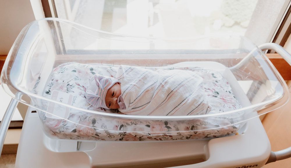 Swaddled newborn baby resting peacefully in a hospital bassinet by the window.