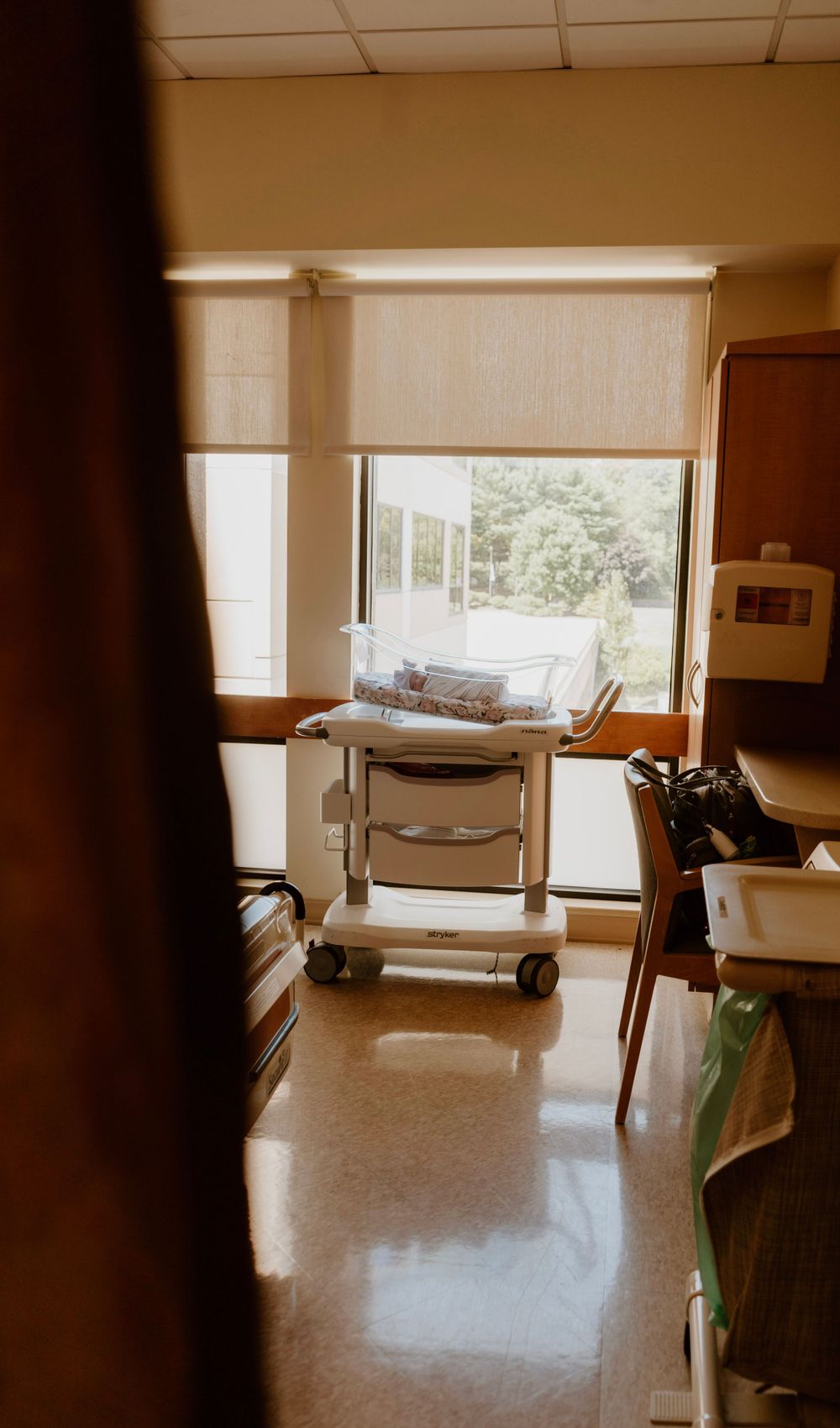 Hospital room with newborn baby in bassinet near a sunlit window.