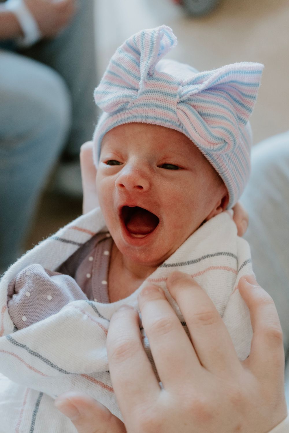 Yawning newborn baby in striped bow hat wrapped in a hospital blanket.