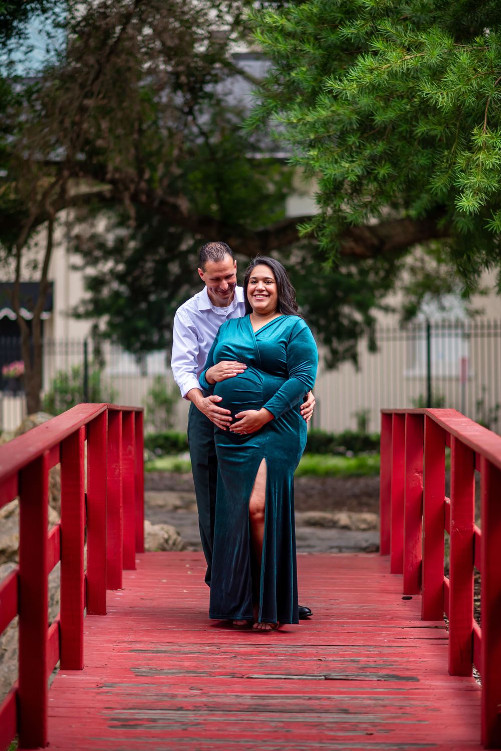 Maternity photo of couple standing on a red bridge in an outdoor garden setting.