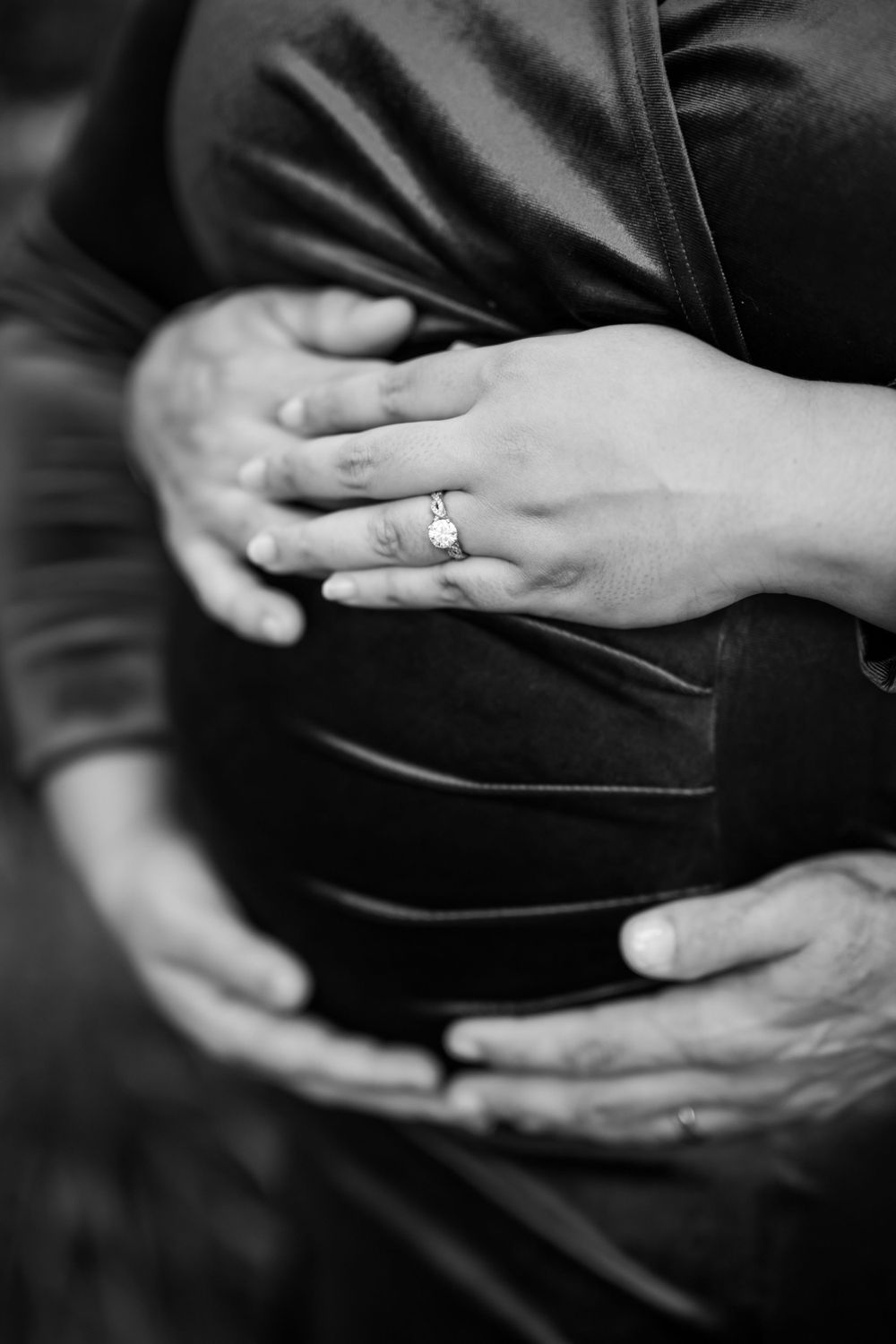 Close-up black and white shot of hands cradling a pregnant belly with engagement ring visible.