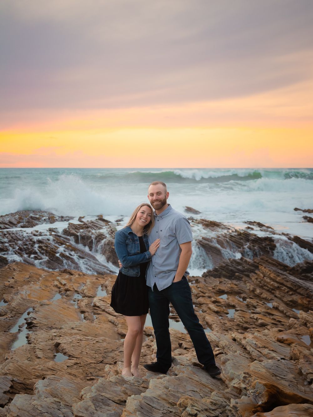 Couple embracing on rocky coastline at sunset with dramatic ocean waves and colorful sky in background.