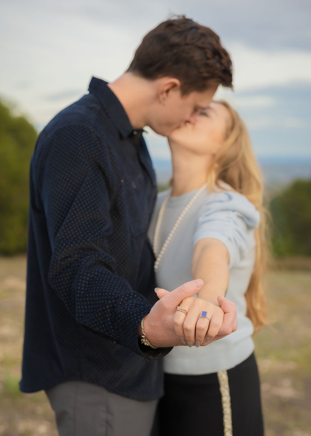 Couple sharing a romantic kiss while showing off an engagement ring outdoors with a blurred natural background.