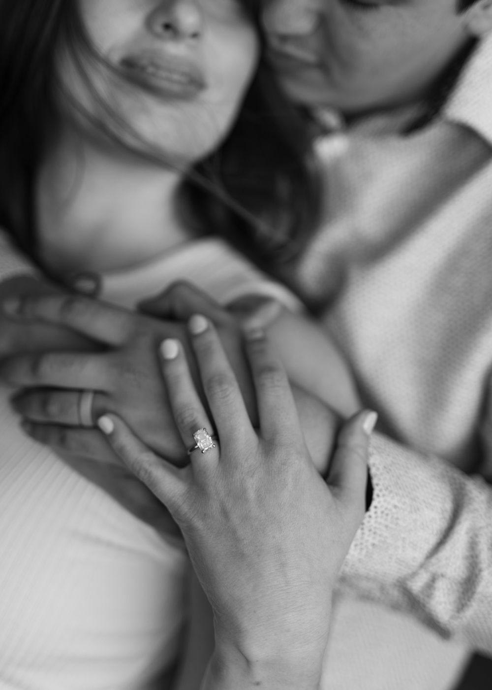 Black and white photo of a couple embracing, showcasing a sparkling diamond engagement ring on the woman's finger.