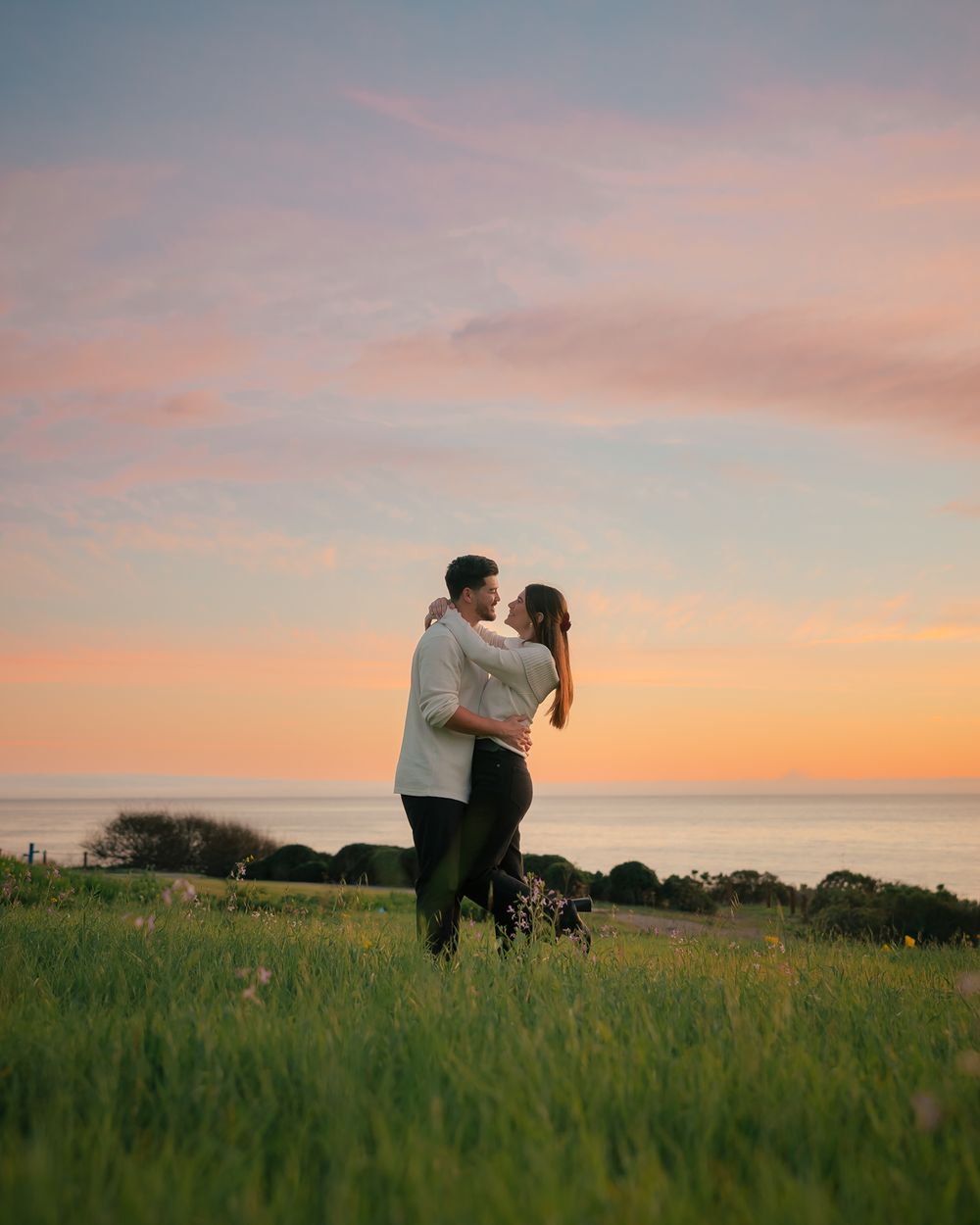A couple shares an intimate moment in a grassy field at sunset with soft pink and orange sky in the background.
