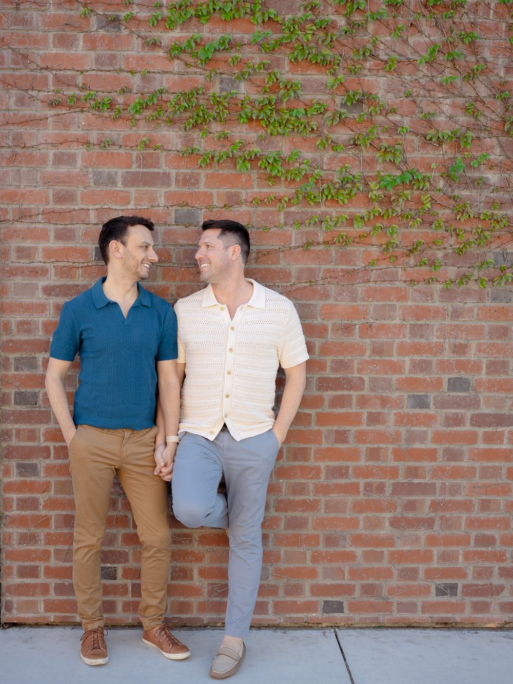 Two men holding hands and smiling at each other against a brick wall with ivy, wearing casual summer outfits.