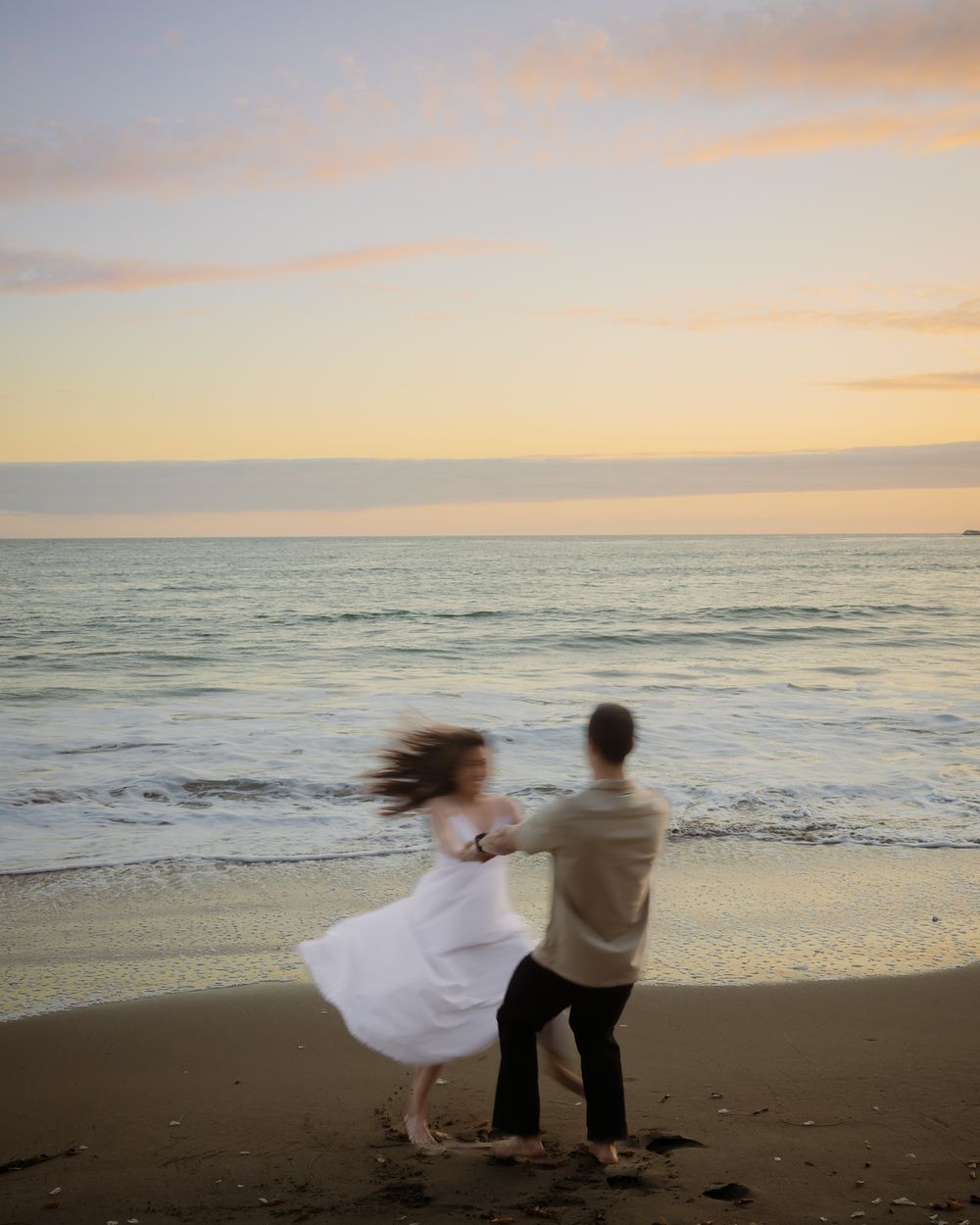 Couple dancing joyfully on a sandy beach at sunset, waves crashing behind them in golden light.