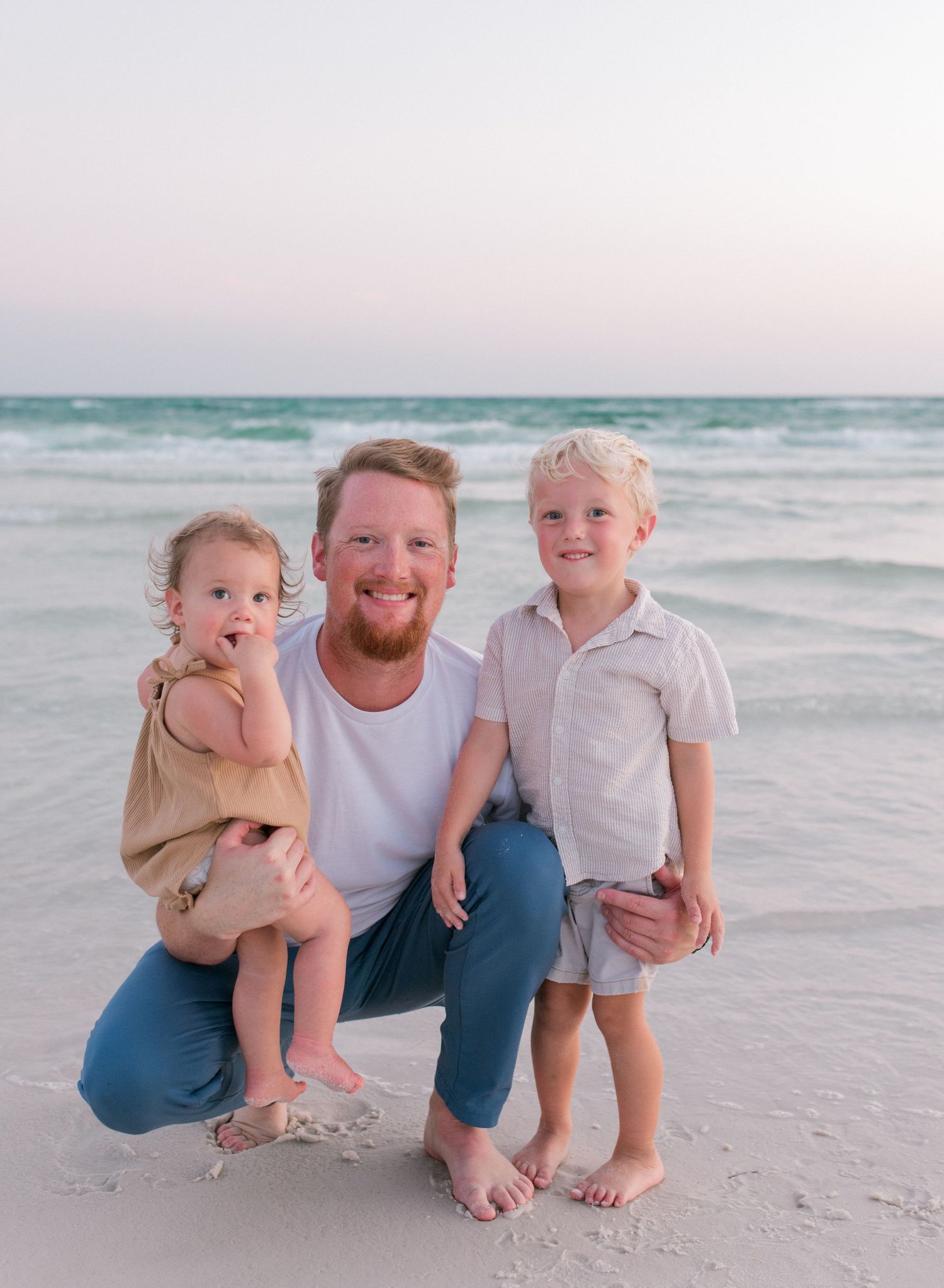A heartwarming beach sunset family portrait with bare feet in the sand and ocean waves in the background.