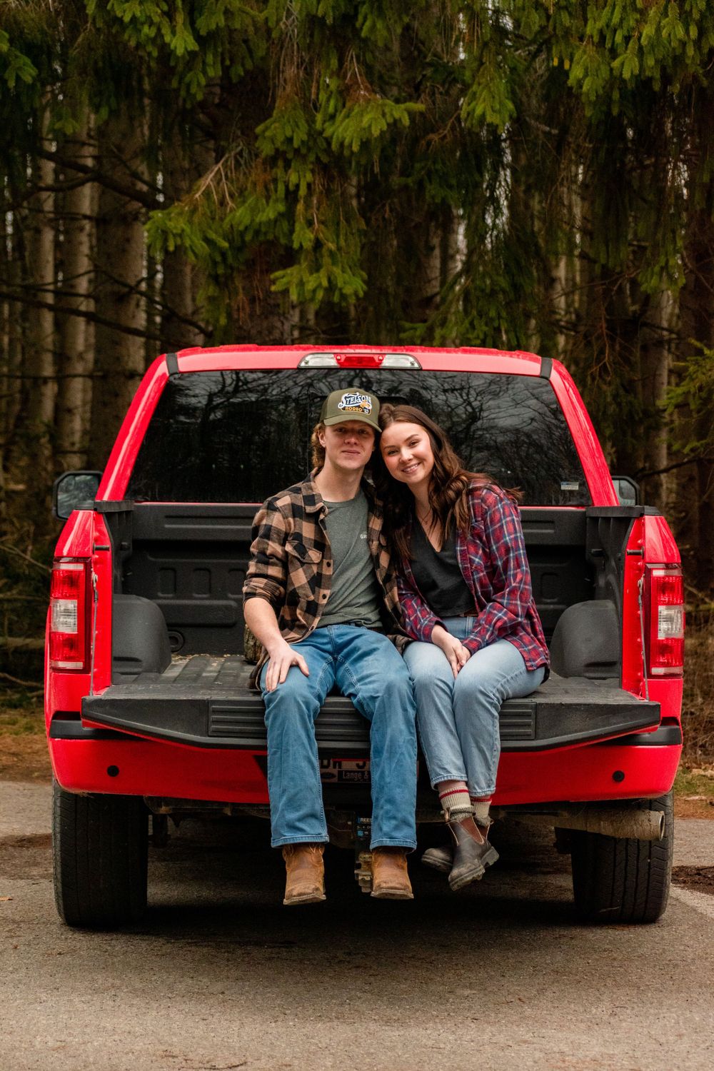 Casual couple sitting together in red pickup truck bed surrounded by pine forest.