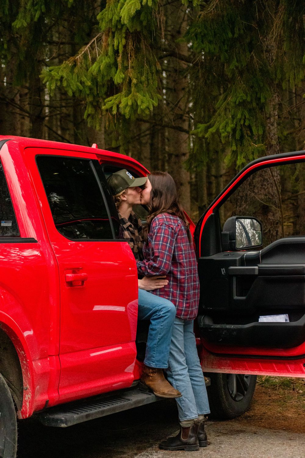 A couple stands beside a bright red pickup truck in a forest setting.