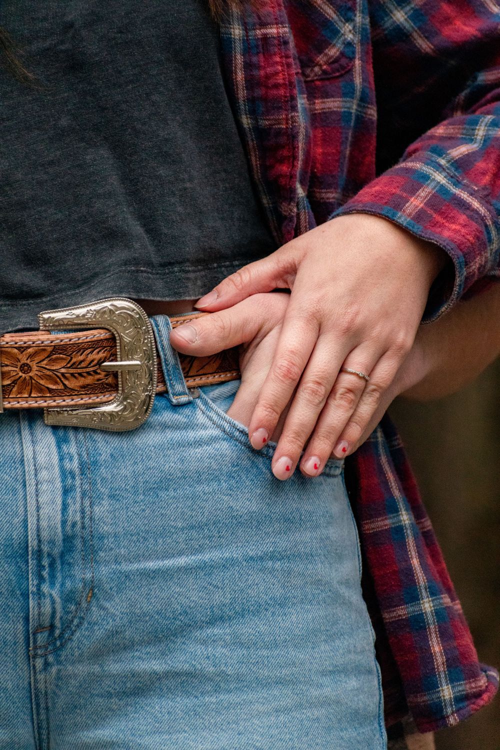 Close-up of hands wearing a ring against jeans and plaid shirt with decorative leather belt.