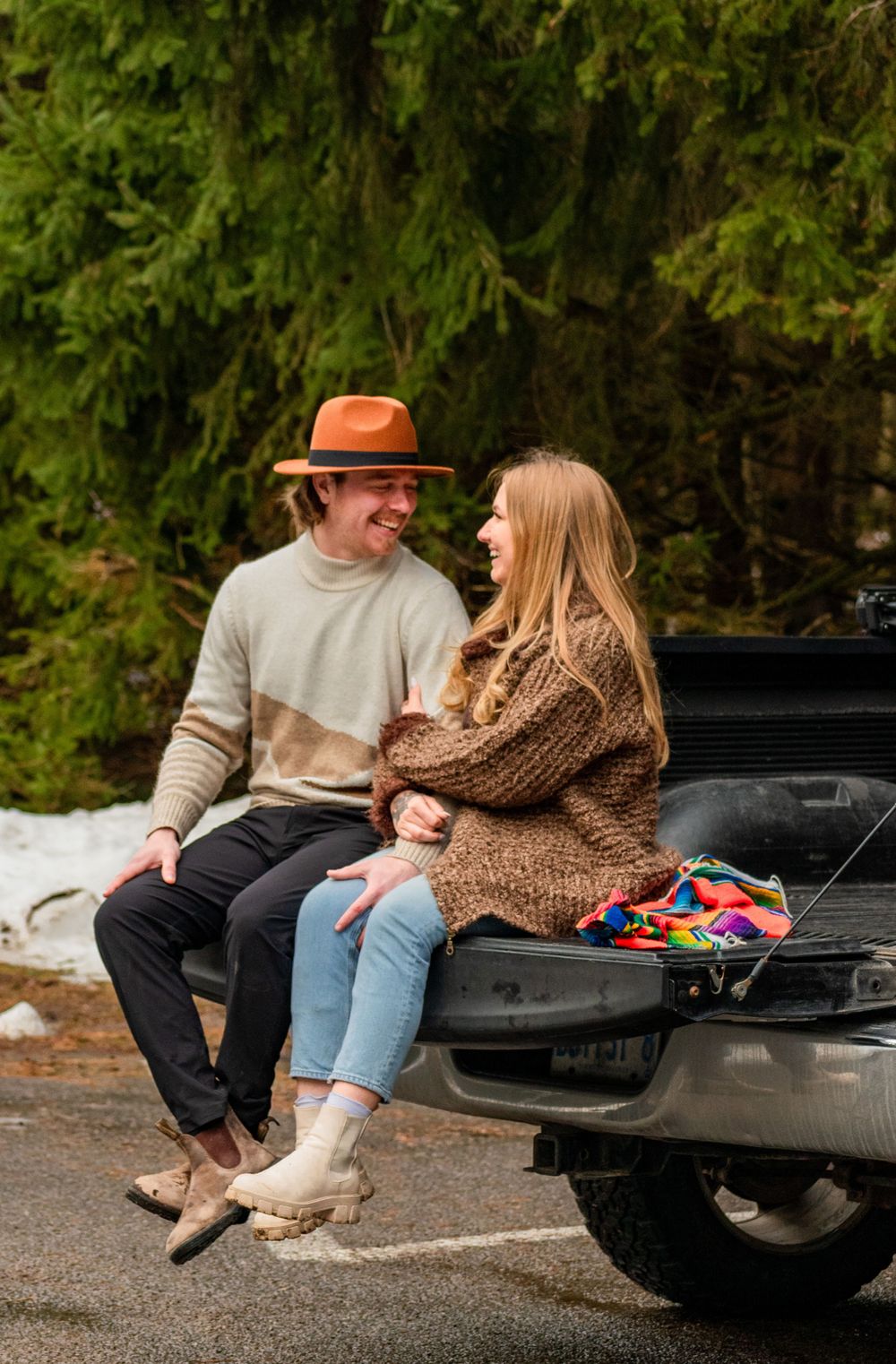 Couple sitting on truck tailgate wearing cozy sweaters in autumn forest setting.