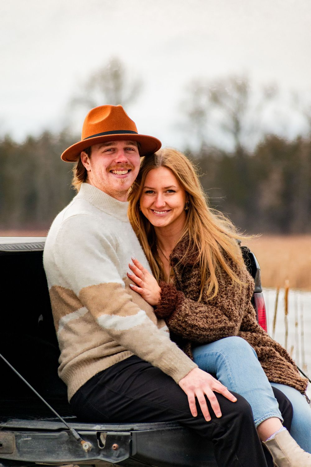 Joyful couple in warm sweaters posing together on truck bed in fall landscape.
