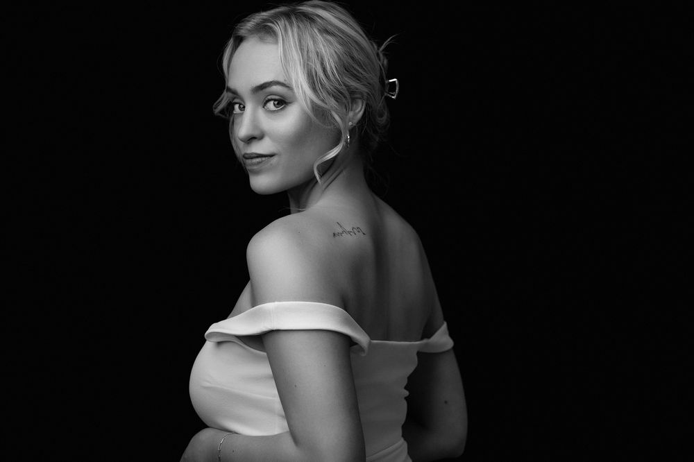 Black and white portrait of a young blonde woman in an off-shoulder dress, looking back over her shoulder.