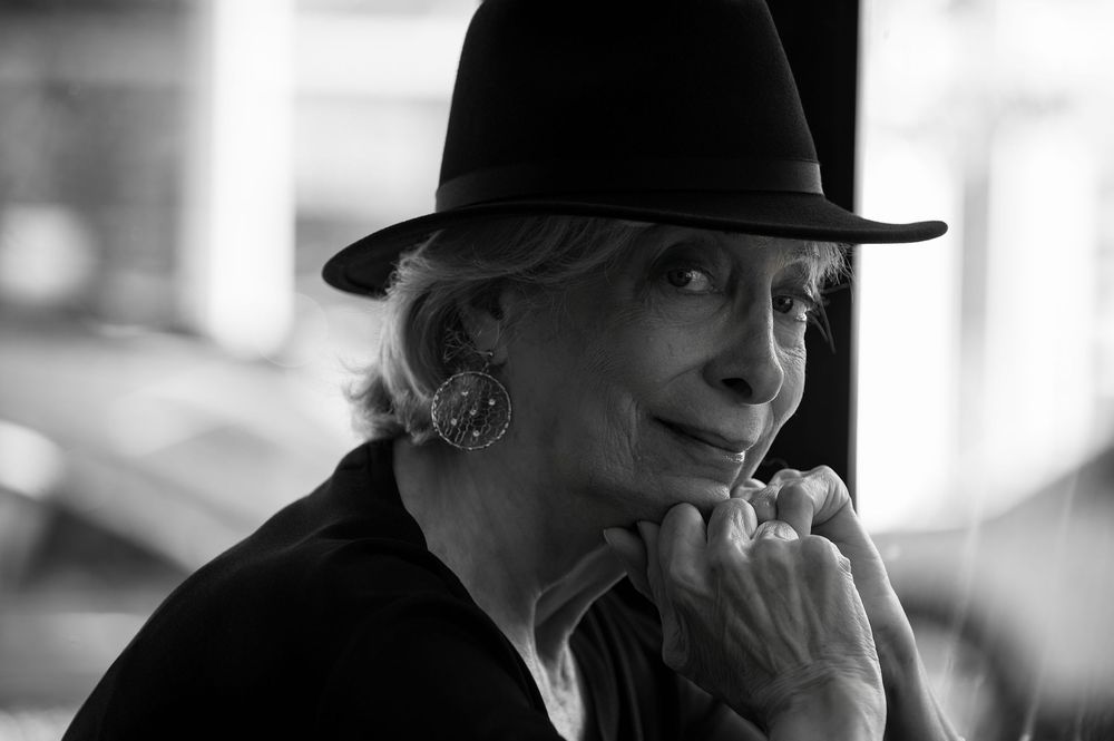 Elderly woman in black hat and earrings posing thoughtfully in black and white portrait photo.