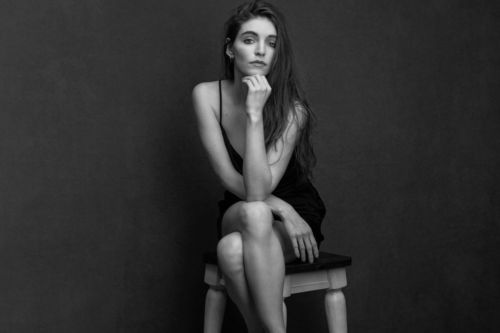 Black and white portrait of a young woman in a black dress sitting on a stool, posing thoughtfully.