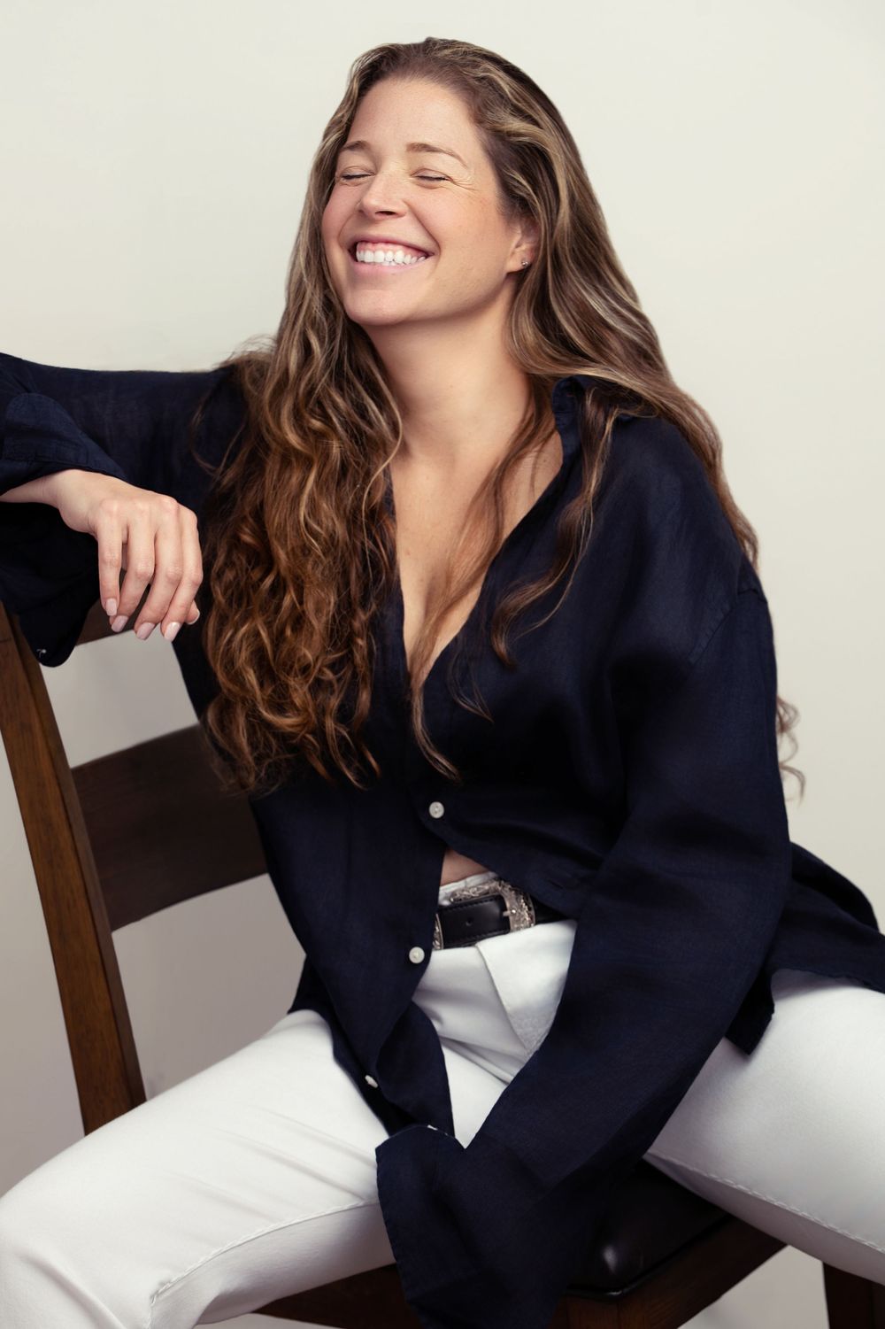 Smiling woman with long wavy hair wearing a navy blouse and white pants, seated on a wooden chair.