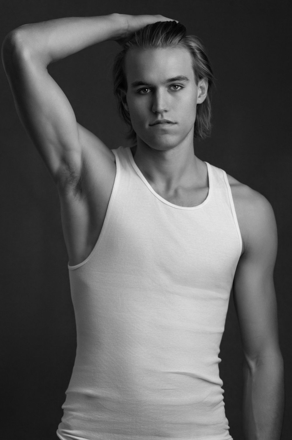 Young male model in white tank top poses confidently against dark background in black and white photo.