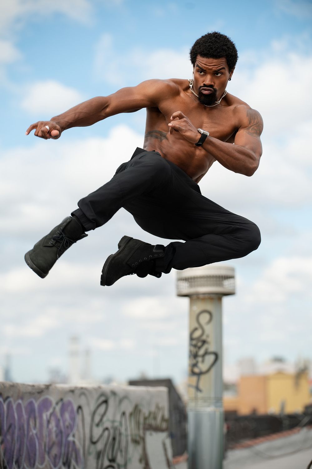 Muscular shirtless Black man performing a martial arts jump kick outdoors against a cloudy sky backdrop.