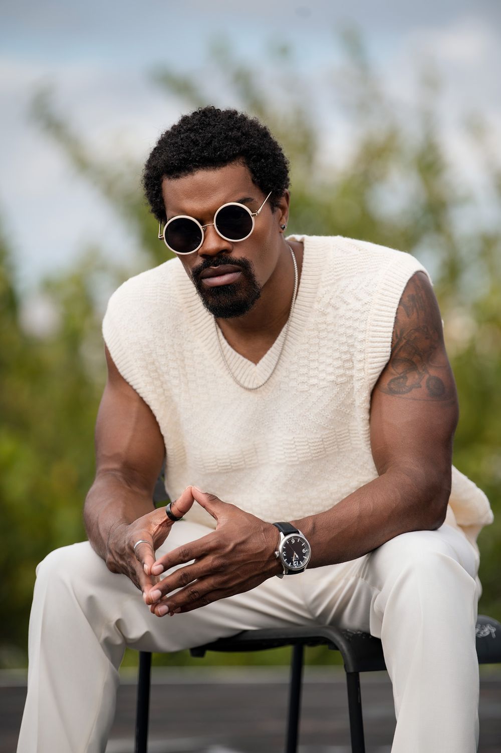 Muscular man in cream knit vest and white pants wearing round sunglasses, sitting outdoors with tattoos visible.