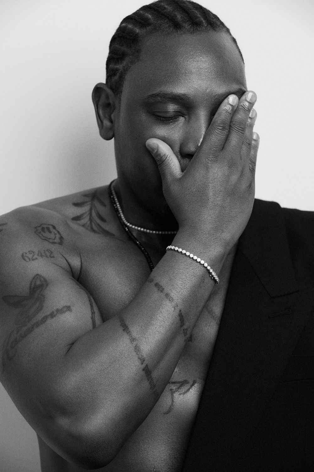 Black and white portrait of a muscular man with braids, tattoos, and a bracelet covering his face with his hand.