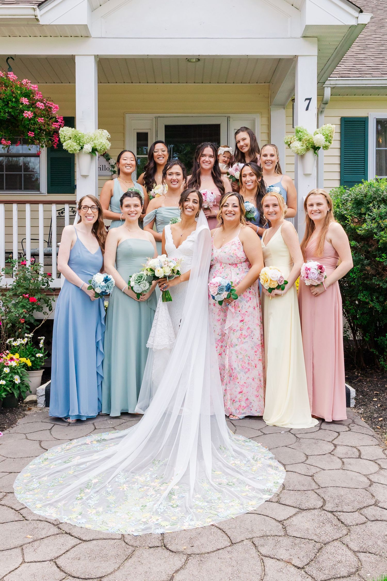 Wedding party poses on stone patio in front of white house with flowing bridal gown and mixed pastel dresses.