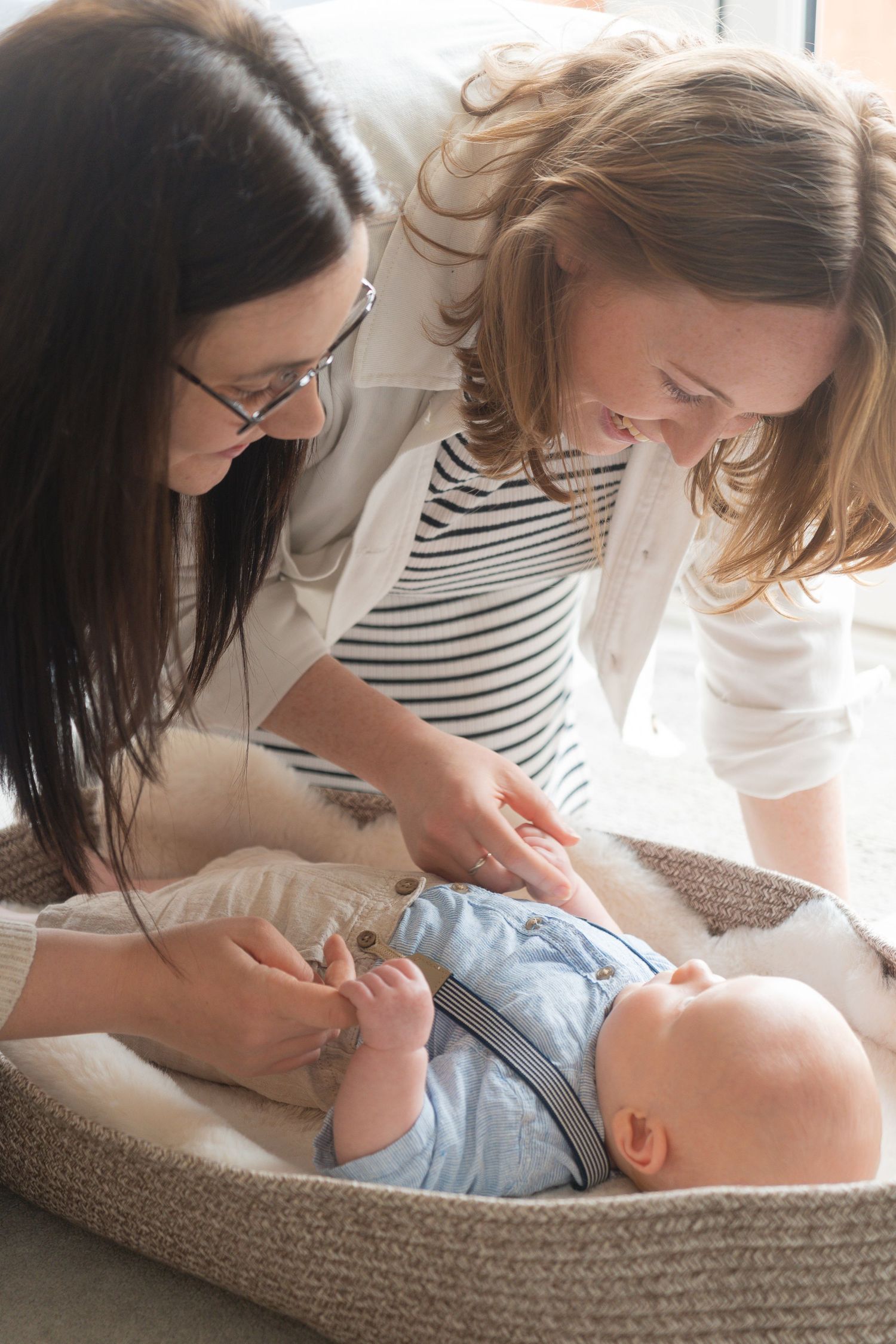 Two mums tickling and smiling at their newborn baby boy who is laying in a basket.