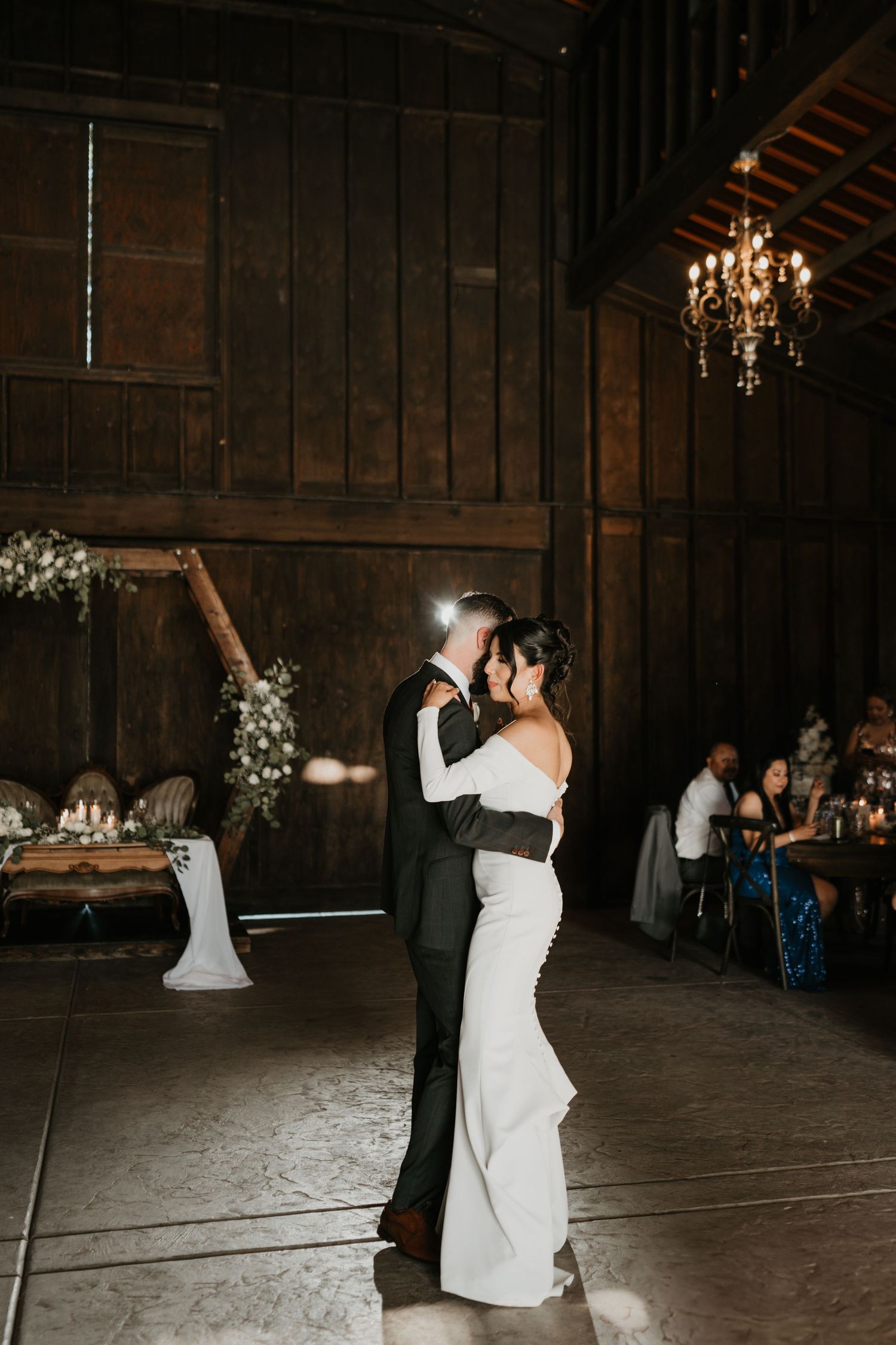 Wedding couple shares first dance under crystal chandelier in rustic barn venue.