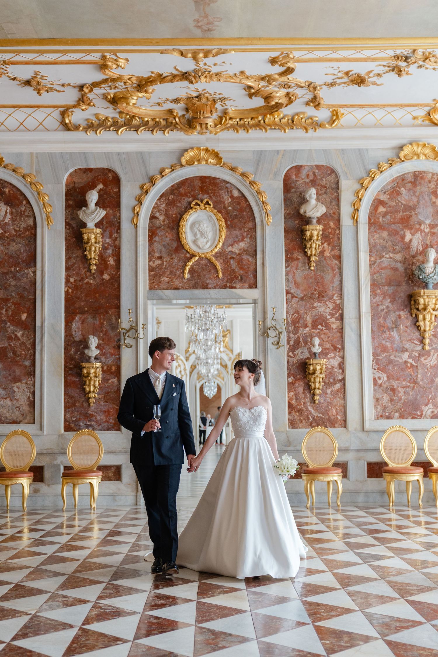 A couple toasts champagne at an elegant wedding reception with crystal glasses and white floral decor in an ornate room.