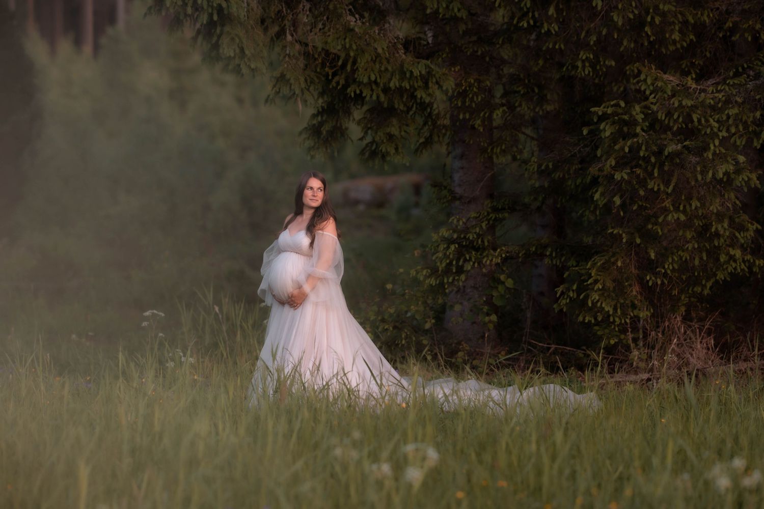 Pregnant woman in elegant white lace gown stands in misty green meadow beside tall evergreens.