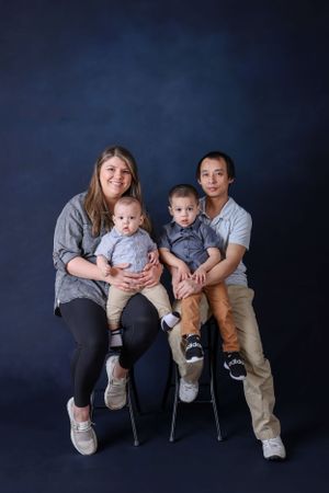 Family portrait of parents with two young sons sitting on chairs against a dark blue backdrop.