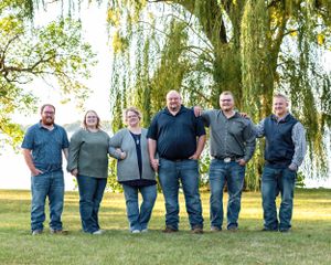 Six adults standing in a relaxed row outdoors near a weeping willow tree by a lake.