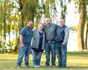 Four family members smiling together on a green lawn near a weeping willow by a lake.