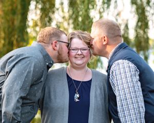 Woman smiling as two men give her a kiss on each cheek outdoors near a willow tree.