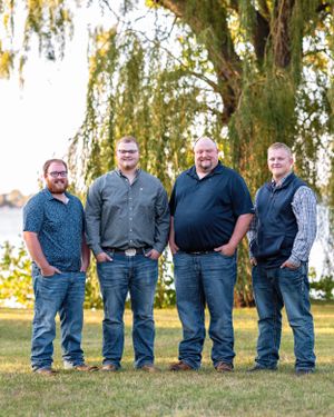 Four men standing together smiling on a green lawn near a weeping willow by a lake.
