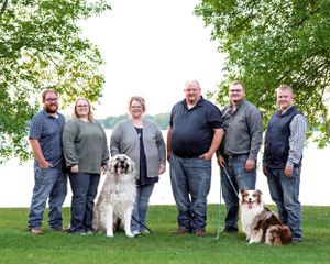 Family group portrait with two dogs outdoors near a lake surrounded by green trees.