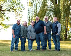 Group of six adults smiling together on a green lawn near a weeping willow by a lake.