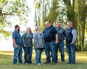 Six adults laughing together outdoors near a weeping willow tree beside a peaceful lake.